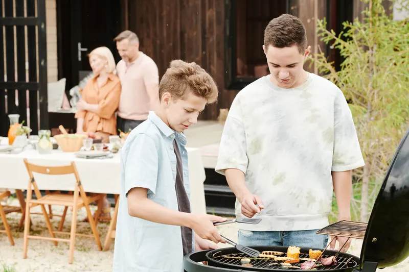 Two young people cooking at a barbecue while their family spends time together outdoors.